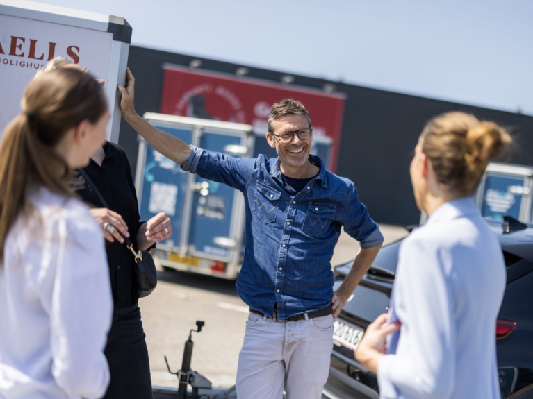 Smiling man talking to a group near branded Freetrailer units, illustrating partnership opportunities and how to become partner in a shared economy concept.